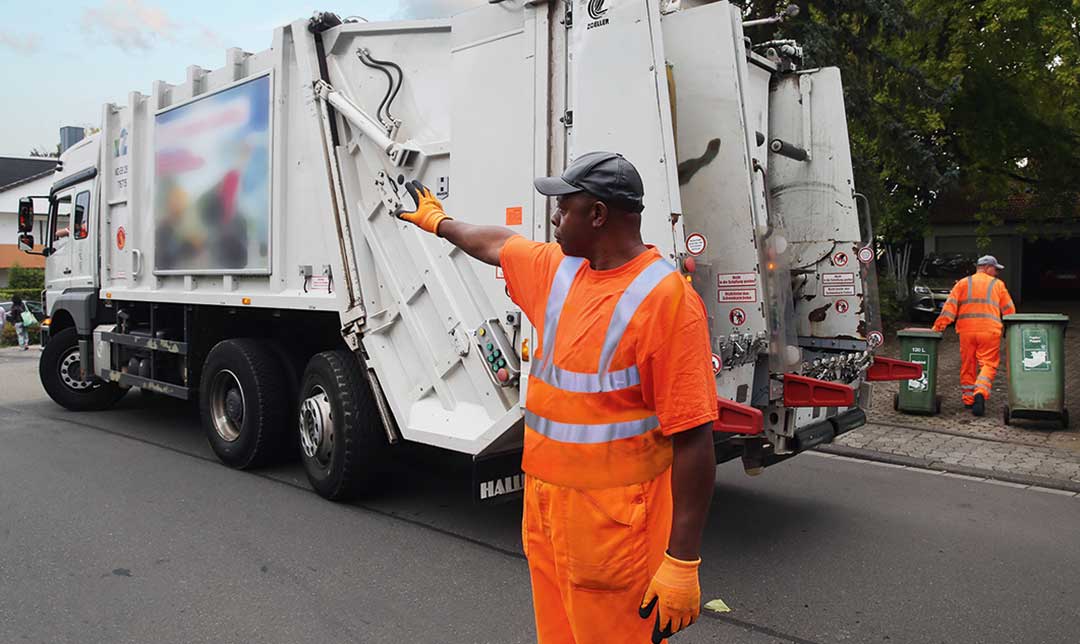 Entsorgungsmitarbeiter steht neben dem Fahrzeug und weist seinen rückwärtsfahrenden Kollegen ein
