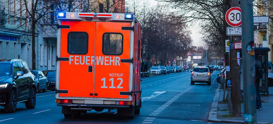 Im Zweifel alarmiert man nach einem Arbeitsunfall den Rettungswagen Blick von hinten auf einen Rettungswagen, der mit Blaulicht eine Straße entlangfährt.