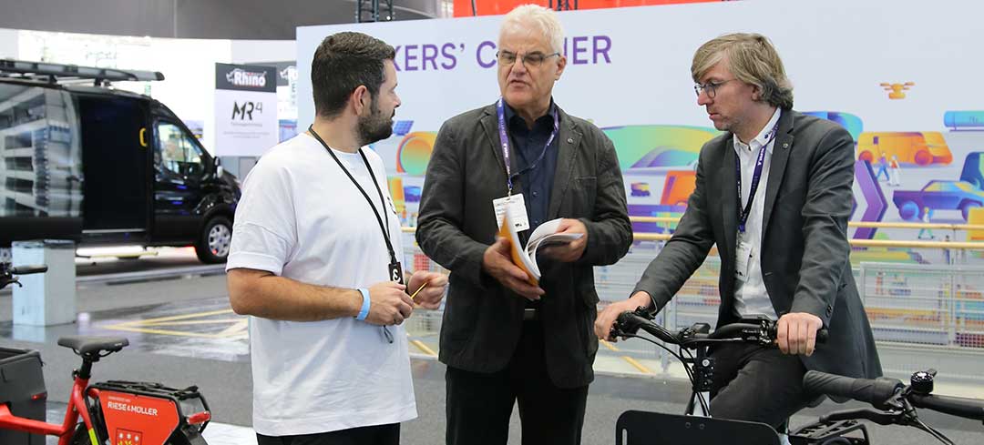 Beratung durch die BG Verkehr auf der IAA Transportation Drei Männer an einem Messestand. Einer von ihnen sitzt auf einem Lastenrad