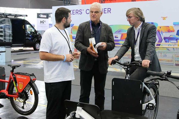 Drei Männer an einem Messestand. Einer von ihnen sitzt auf einem Lastenrad