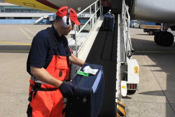 Mann mit Gehörschutz lädt Koffer auf ein Förderband, das in einen Flugzeugladeraum führt