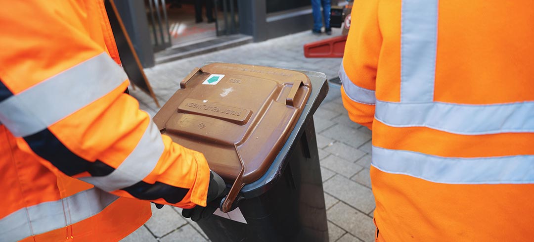 Gefährdungen bei der Sammlung von Bioabfällen Mitarbeiter eines Entsorgungsbetriebs bringen eine braune Bio-Tonne zu ihrem Fahrzeug