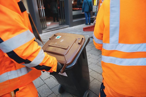 Mitarbeiter eines Entsorgungsbetriebs bringen eine braune Bio-Tonne zu ihrem Fahrzeug