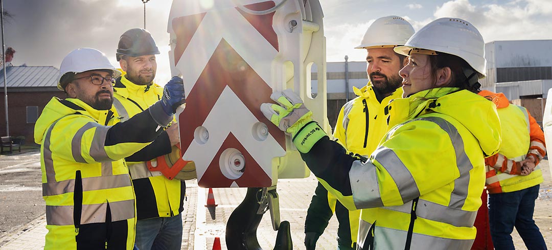 Aufsichtspersonen der BG Verkehr bilden sich regelmäßig weiter Mitarbeiter stehen im Kreis um den Kranhaken eines Autokrans