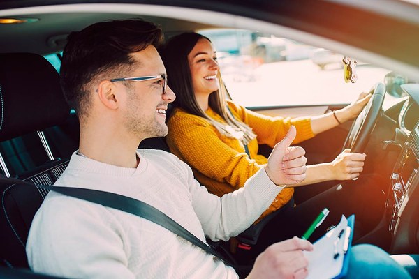 Fahrlehrer zeigt der Fahrschülerin am Steuer lächelnd einen "Daumen hoch!"