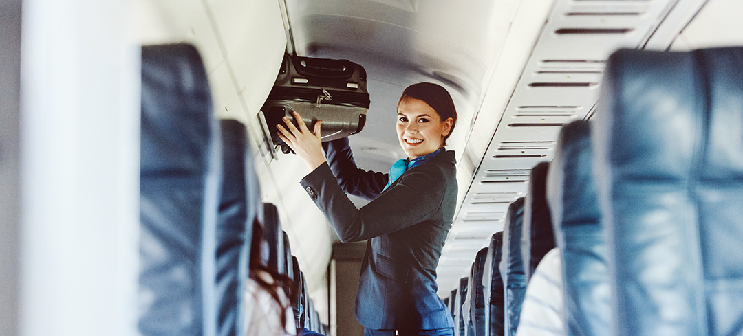 Foto einer lächelndne Flugbegleiterin, die einen Koffer in das obere Gepächfach verräumt