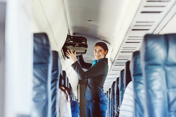 Foto einer lächelndne Flugbegleiterin, die einen Koffer in das obere Gepächfach verräumt