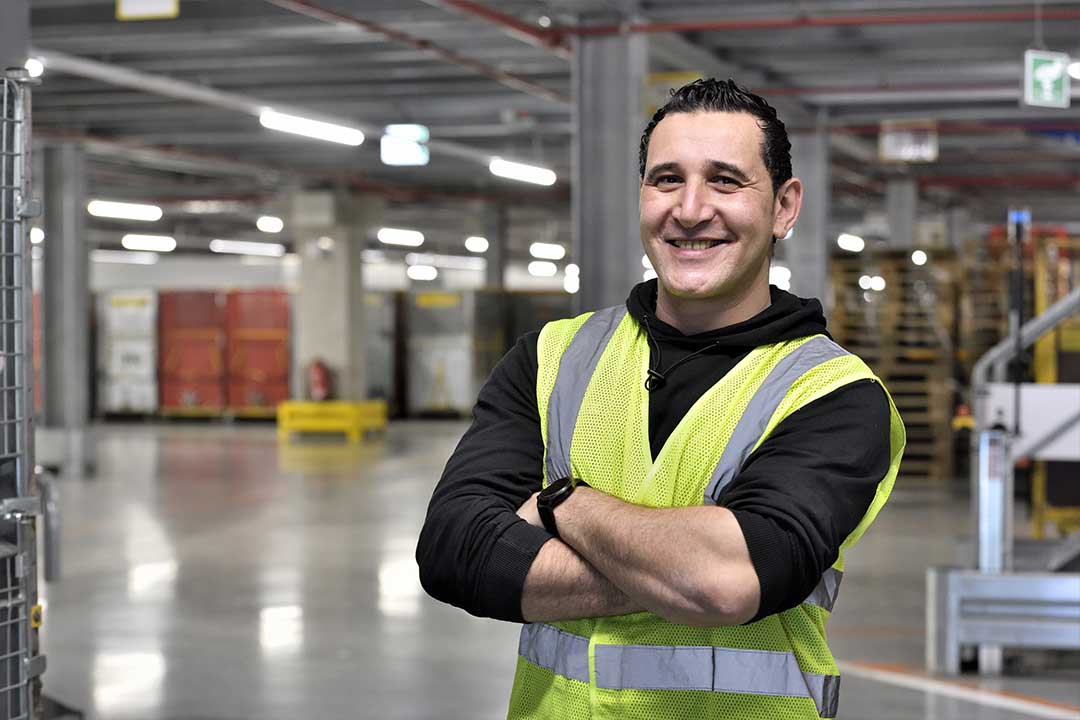 Die Beschäftigten profitieren vom High-Tech im Paketzentrum Ein Mann steht im Paketzentrum mit schwarzen Haaren und gelber Warnweste lächelt mit verschränkten Armen in die Kamera.