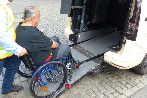 Ein Mann sitzt in einem Rollstuhl und wird eine Rampe hinauf in einen kleinen Transporter geschoben.