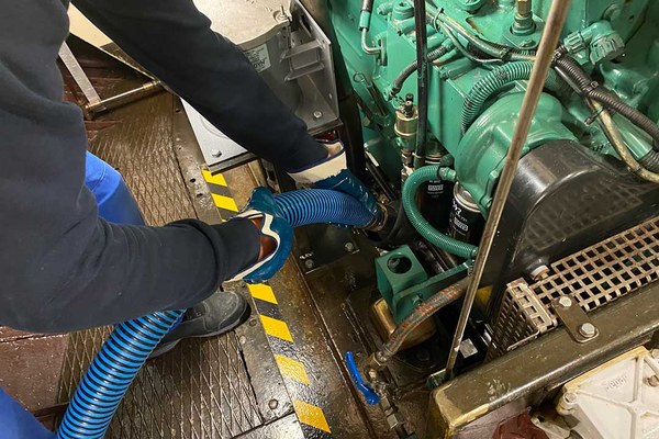 Zwei Hände mit Handschuhen halten einen Schlauch fest, der in den Motorraum eines Schiffes führt.