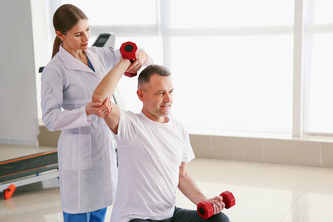 Physiotherapeutin hilft einem Patienten bei einer Rückenübung mit Hanteln