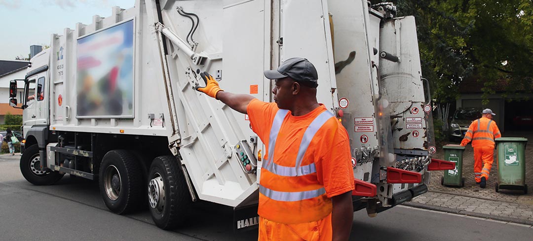 Entsorgungsmitarbeiter steht neben dem Fahrzeug und weist seinen rückwärtsfahrenden Kollegen ein
